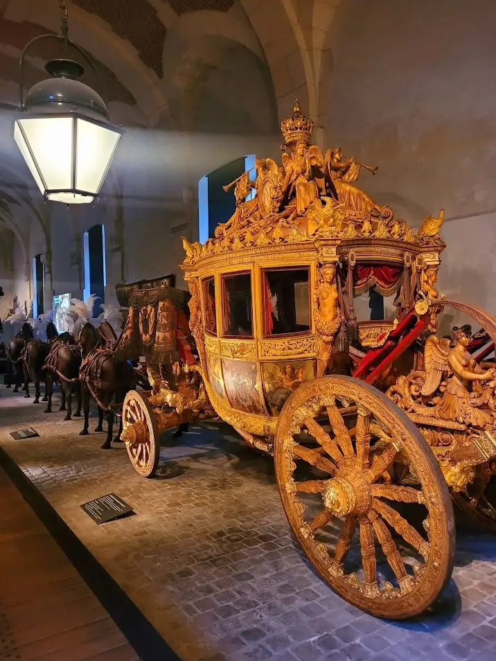 Side view of Napoleon’s coronation carriage in the Coach Gallery at the Palace of Versailles, richly gilded with mythological sculptures, crowned imperial emblems, and painted panels, drawn by a full team of black model horses under the vaulted ceiling of the historic stables.