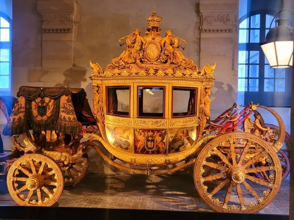 Magnificently ornate imperial carriage in the Coach Gallery at the Palace of Versailles, adorned with intricate gold detailing, sculpted figures, painted mythological scenes, and imperial emblems, including a large crowned “N” crest on top, flanked by figures representing Napoleon’s reign.