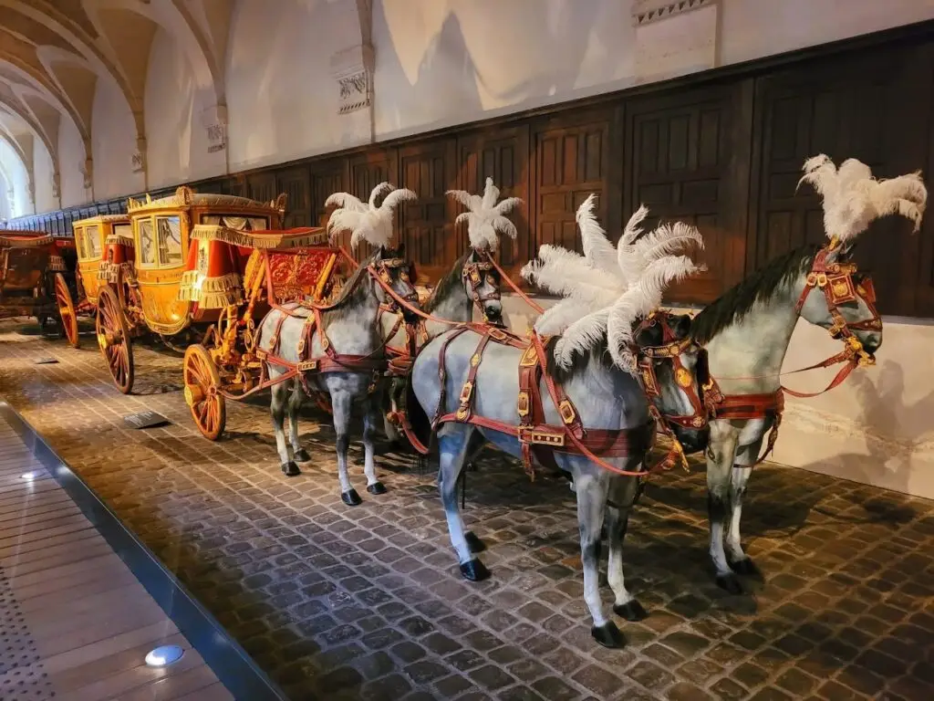 Row of opulent royal carriages displayed in the Coach Gallery at the Palace of Versailles, each elaborately decorated with gilded panels and red velvet canopies, accompanied by a team of model horses adorned with red harnesses and white feather plumes, arranged along a cobblestone floor under vaulted ceilings.