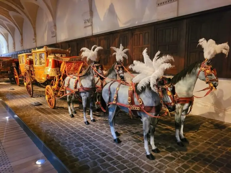 Row of opulent royal carriages displayed in the Coach Gallery at the Palace of Versailles, each elaborately decorated with gilded panels and red velvet canopies, accompanied by a team of model horses adorned with red harnesses and white feather plumes, arranged along a cobblestone floor under vaulted ceilings.