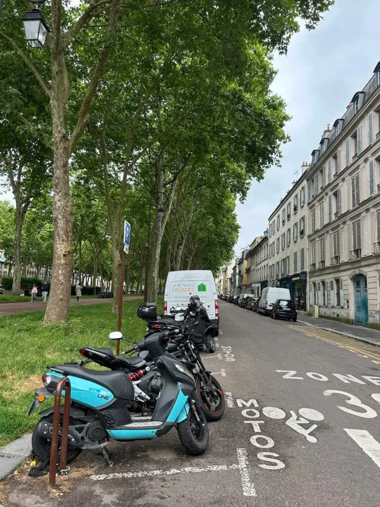 Tree-lined street in Versailles with motorcycles parked in a designated 'motos' zone and classic French apartment buildings on the right-hand side.