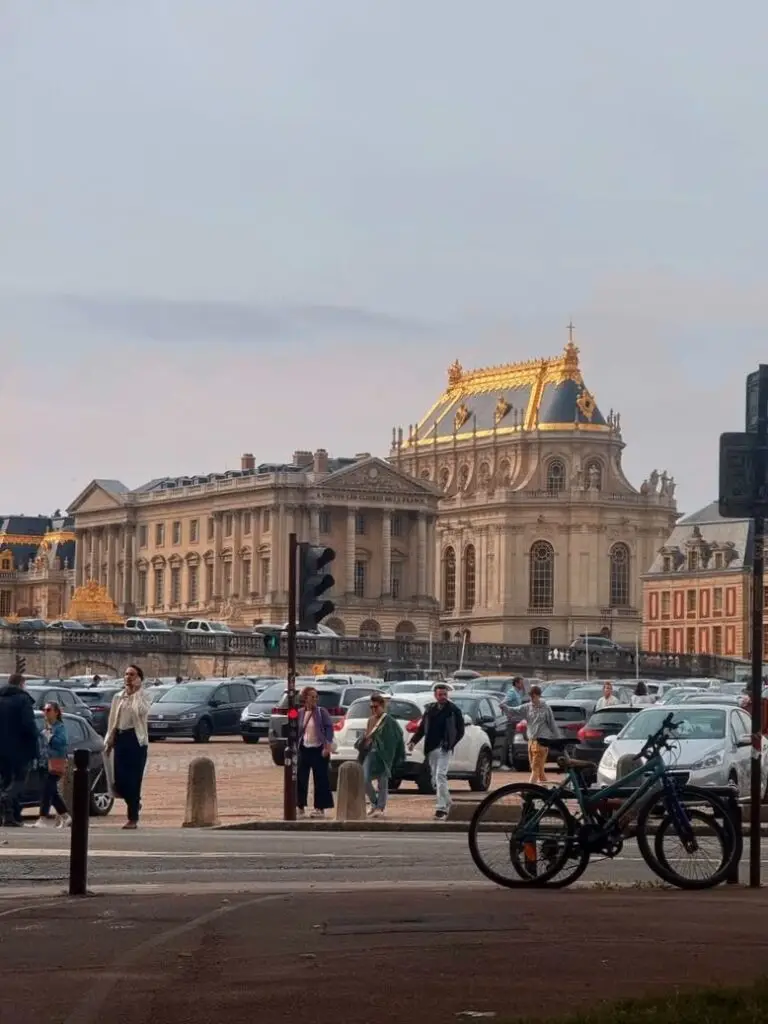 Visitors walking past parked cars in Place d'Armes, directly in front of the Palace of Versailles, with the golden roof of the Royal Chapel illuminated in the background.