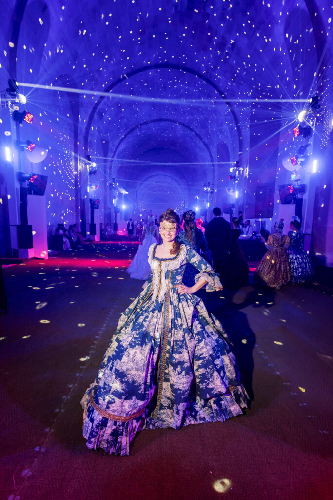 Posing confidently beneath a starry light show at the Grand Bal Masqué 2025 in Versailles, I wear a blue toile gown and golden mask, surrounded by guests in period dress inside the palace’s dazzling ballroom.