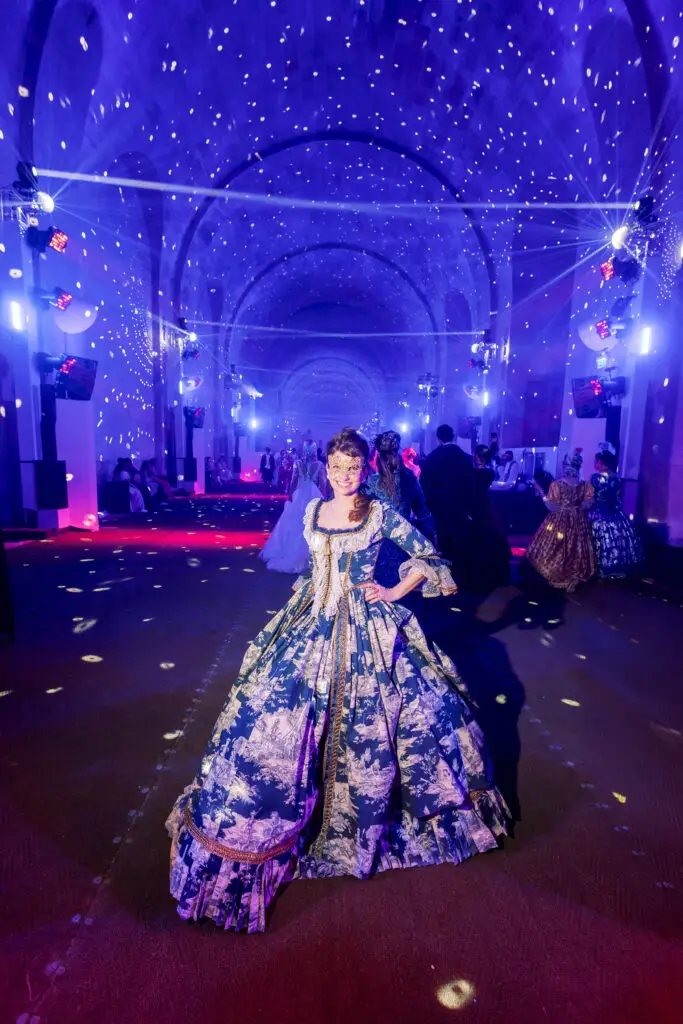 Posing confidently beneath a starry light show at the Grand Bal Masqué 2025 in Versailles, I wear a blue toile gown and golden mask, surrounded by guests in period dress inside the palace’s dazzling ballroom.