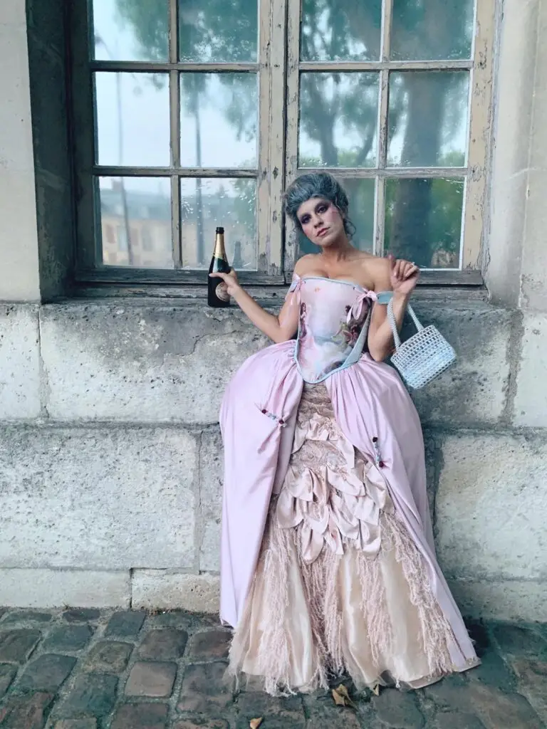 Tara Lengyel dressed in an elaborate, Rococo-style costume reminiscent of Marie Antoinette leans casually against a stone ledge in front of an old window. They wear a voluminous pink and cream gown with ornate detailing, powdered hair styled high, dramatic makeup, and hold a bottle of champagne in one hand and a cigarette in the other. A small light blue handbag hangs from their arm. The setting appears to be a historic stone building with cobblestone ground.