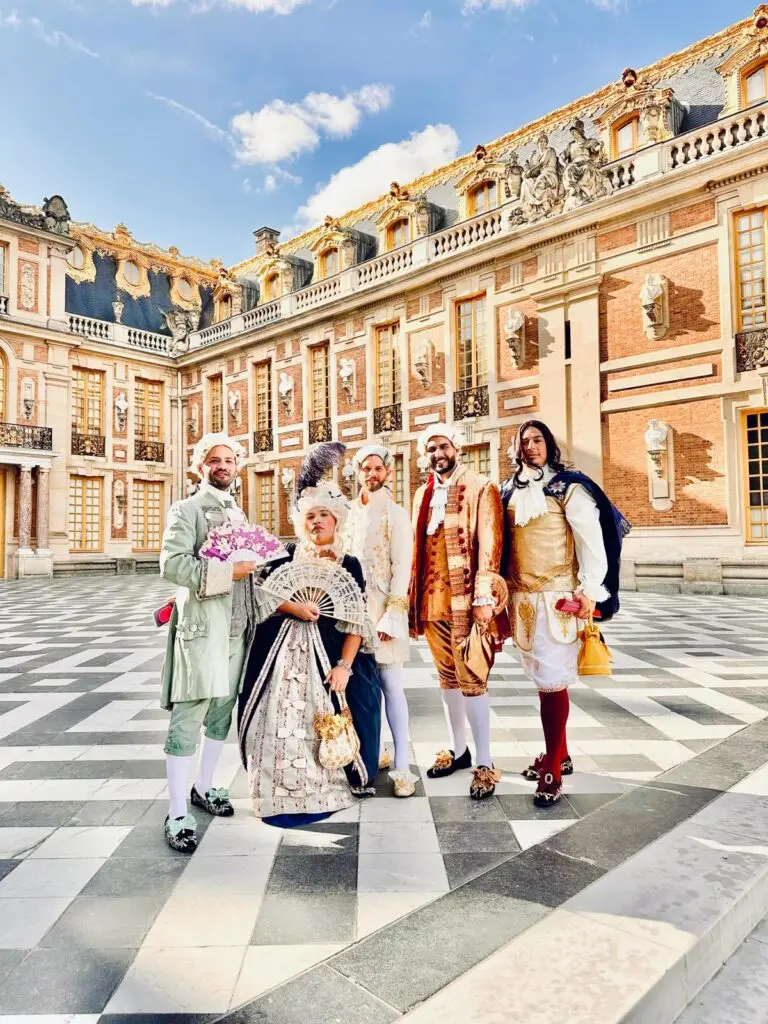 Group of guests in elaborate baroque costumes pose in the marble courtyard of the Palace of Versailles under a bright sky—perfectly capturing the grandeur of the Versailles masquerade experience.
